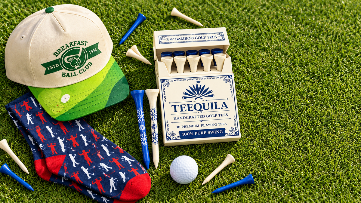 Funny golf gifts flat-lay: Teequila golf tee box, Breakfast Ball Club golf hat and golf socks on green turf
